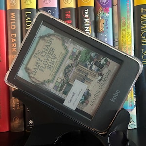 Op de afbeelding: Een zwarte Kobo e-reader in een doorzichtige beschermhoes, rustend op een zwarte plastic standaard. Het scherm toont de boekomslag van The Daisy Chain Flower Shop door Laurie Gilmore, met een statusbalk die Sleeping en 55% read aangeeft.