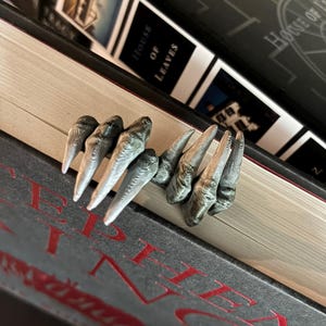 May include: A gray, textured bookmark shaped like a pair of clawed hands with long, pointed fingers. The bookmark is positioned between the pages of a closed book, with the fingers extending outward from the edge of the book pages.
