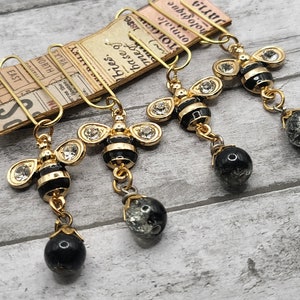 May include: Four gold bee charms with black and white stripes and clear rhinestones. Each charm is attached to a black bead and a gold clip.