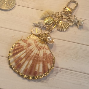May include: A gold-toned keychain featuring a large, fan-shaped seashell with a gold-painted edge. The keychain is adorned with various small seashells, a starfish charm with a pearl, and a circular charm with the letter "Z".