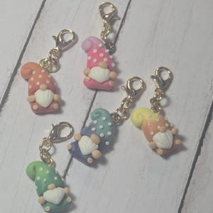 May include: A collection of colorful gnome charms with gold-colored clasps. Each gnome has a pointed hat with polka dots, a white heart-shaped beard, and small beige arms and legs. The gnome hats are in shades of pink, orange, yellow, green, and blue.