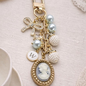 May include: Gold-toned keychain with a bow, pearls, and a cameo pendant. The keychain features a lobster clasp, a bow embellished with rhinestones, light blue pearls, and a cameo pendant with a woman's profile.
