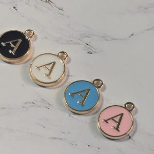 May include: Four round charms with the letter "A". The charms are black, white, blue, and pink with gold-colored trim. Each charm has a small loop at the top for attaching to a necklace or bracelet. Jewelry making supplies.