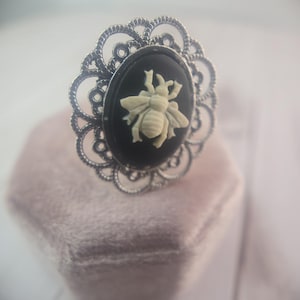 May include: A silver-toned ring with an oval black cameo featuring a cream-colored bee. The cameo is set within a decorative, openwork frame. The ring is a piece of jewelry.