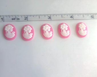 May include: Five oval-shaped pink cameo cabochons with white relief portraits of women. The cabochons are arranged in a row, next to a ruler marked in inches. These cabochons are ideal for jewelry making or craft projects.
