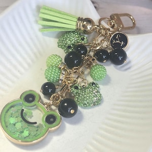 May include: A whimsical keychain featuring a green frog charm with a smiling face and a shaker design filled with glitter. The keychain also includes a green tassel, black beads, and small green frog charms, all connected with gold-tone hardware.