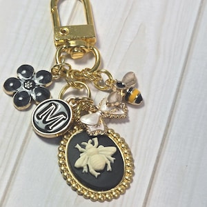 May include: Gold-toned keychain with multiple charms. Charms include a black and white flower, a round charm with the letter "M", a bee, and a cameo with a bee design. The keychain has a lobster clasp.