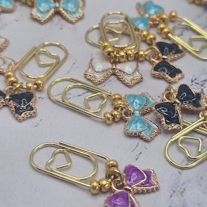 May include: A collection of gold-colored paper clips, each adorned with a small, decorative bow charm. The bows come in various colors, including light blue, white, black, and purple, with gold-colored trim. The charms are attached with small gold beads.