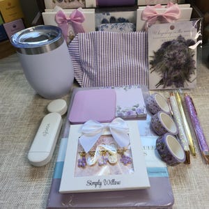 May include: A collection of stationery and lifestyle items including a light purple tumbler, a striped fabric pouch, a notebook, floral washi tapes, glittery pens, a white Vavofo device, and several packaged sets of decorative paper clips and stickers.