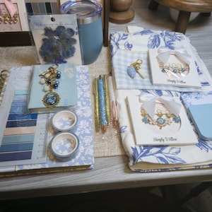 May include: A collection of stationery and craft items including a blue tumbler, a floral notebook, two rolls of washi tape, three glitter pens, and two sets of Simply Willow brand decorative paper clips with white bows on a blue and white floral fabric.