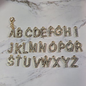 May include: A collection of gold-colored alphabet charms, each encrusted with small, clear crystals. The letters A through Z are displayed, along with a clasp. These charms are suitable for jewelry making or personalized crafts.