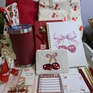 May include: A collection of stationery and accessories including a red tumbler, a cream cherry-print tote bag, a spiral notebook with a pink bow and cherry illustration, a beaded cherry coin purse, two notepads, pens, and various cherry-themed charms.