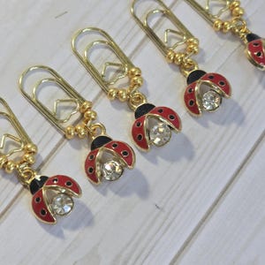 May include: Gold-toned paper clips with ladybug charms. Each ladybug charm features a red enamel body with black spots and a clear crystal center. The ladybug charms are attached to the paper clips with gold-colored beads.