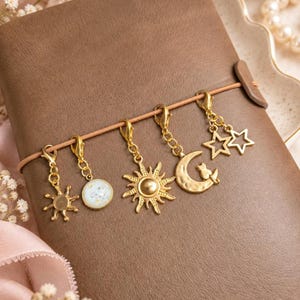 May include: A brown notebook with an elastic band featuring five gold-toned metal charms attached by lobster clasps. The charms include a small sun, a round white speckled disc, a detailed sun, a crescent moon with a cat, and two stars.