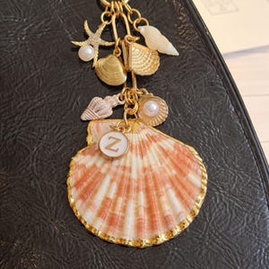 May include: A gold-toned keychain featuring various seashell charms, including a starfish with a pearl, a scallop shell, and a large, striped seashell pendant with a gold border and the letter "Z".