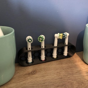May include: A black plastic toothbrush head holder with four toothbrush heads. Two of the heads are green and white, one is yellow and white, and one is blue and white. The holder is sitting on a wooden surface.