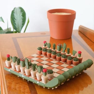 K&ouml;nnte beinhalten: Ein Schachspiel mit Kakteen-Motiv, bei dem Miniatur-Kakteen in T&ouml;pfen als Schachfiguren dienen. Das Schachbrett hat ein braun-cremefarbenes Schachmuster. Im Hintergrund steht ein Terrakotta-Topf. Das Schachspiel steht auf einer Holzoberfl&auml;che.