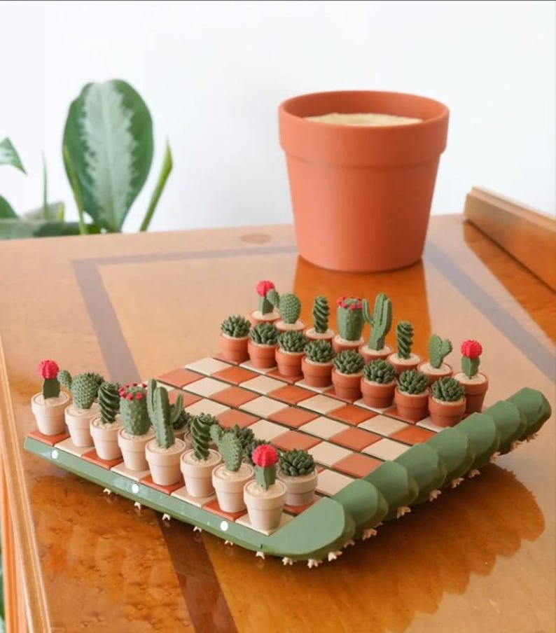 K&ouml;nnte beinhalten: Ein Schachspiel mit Kakteen-Motiv, bei dem Miniatur-Kakteen in T&ouml;pfen als Schachfiguren dienen. Das Schachbrett hat ein braun-cremefarbenes Schachmuster. Im Hintergrund steht ein Terrakotta-Topf. Das Schachspiel steht auf einer Holzoberfl&auml;che.