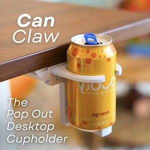 May include: A white plastic "Can Claw" desktop cupholder attached to a wooden table, holding a yellow can of soda with orange spots. The can has a blue pull tab. Text on the image reads "Can Claw" and "The Pop Out Desktop Cupholder."