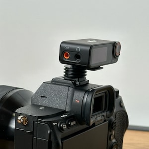 Puede incluir: Una cámara negra con un micrófono externo negro conectado a la parte superior. El micrófono tiene una pequeña pantalla y dos botones en el lateral. La cámara tiene una lente negra y una correa negra.