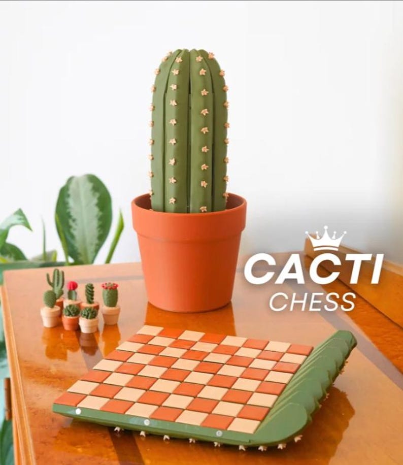 K&ouml;nnte beinhalten: Ein Schachspiel mit Kaktus-Motiv, mit einem gr&uuml;n-orangen Schachbrett und einer gro&szlig;en Kaktusfigur in einem Terrakotta-Topf. Das Set enth&auml;lt Miniatur-Kaktusfiguren und den Text "CACTI CHESS".