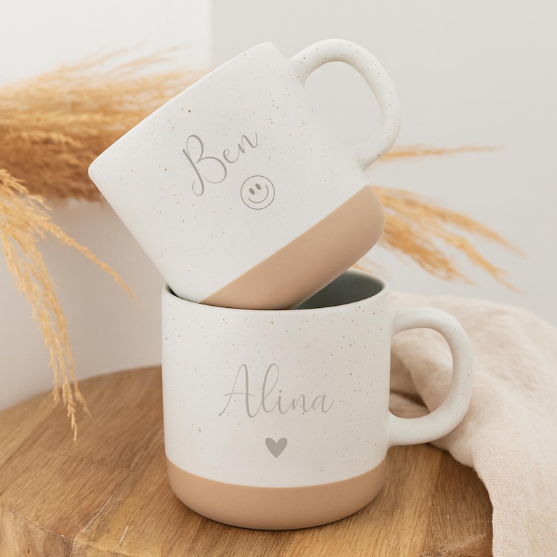 May include: Two white speckled ceramic mugs with tan bottoms and handles are stacked. The top mug has the name "Ben" and a smiley face. The bottom mug has the name "Alina" and a heart symbol.