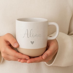 May include: A white speckled ceramic mug with a beige base, featuring the name "Alina" in cursive script and a small heart. The mug has a large handle and is held in both hands.