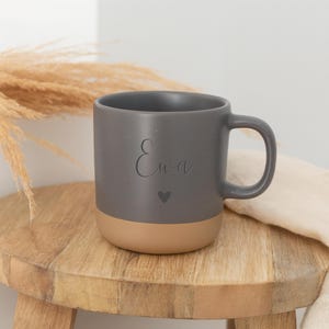 May include: A gray ceramic mug with a tan base, featuring the name "Ewa" and a heart symbol. The mug is on a small wooden stool, with dried plants in the background.