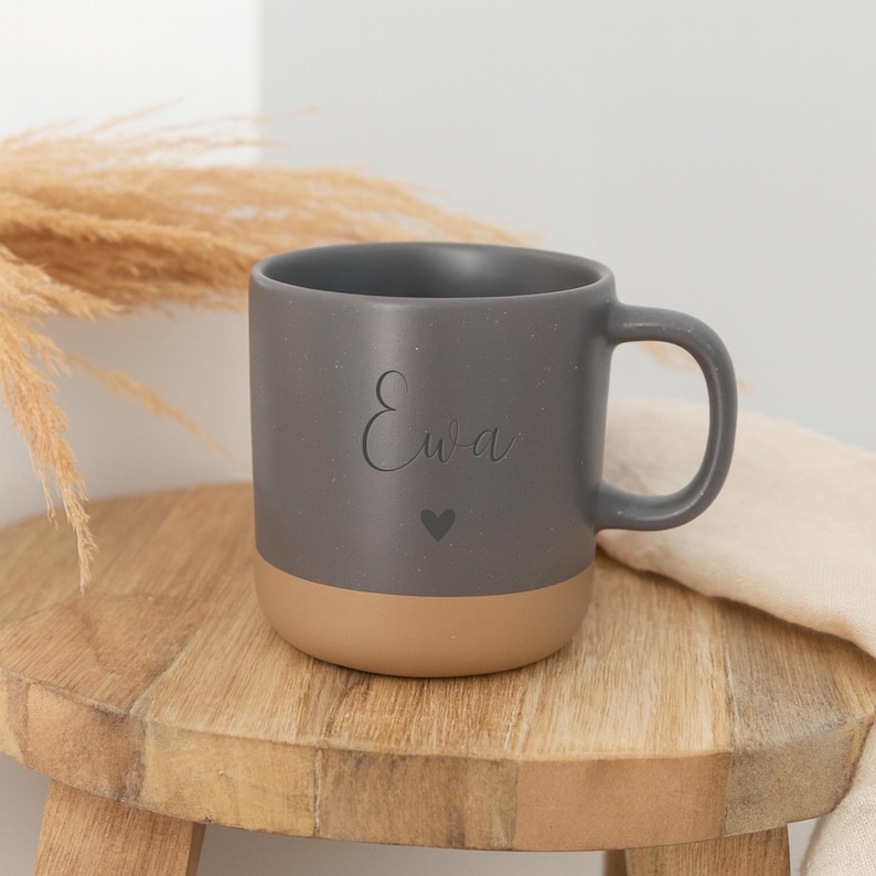 May include: A gray ceramic mug with a tan base, featuring the name "Ewa" and a heart symbol. The mug is on a small wooden stool, with dried plants in the background.