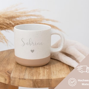 May include: A speckled ceramic mug with the name "Sabrina" in cursive and a small heart. The bottom half of the mug is a solid tan color. The mug sits on a wooden stool with a neutral-colored cloth and dried plants in the background.