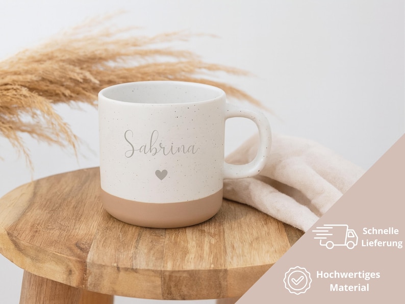 May include: A speckled ceramic mug with the name "Sabrina" in cursive and a small heart. The bottom half of the mug is a solid tan color. The mug sits on a wooden stool with a neutral-colored cloth and dried plants in the background.