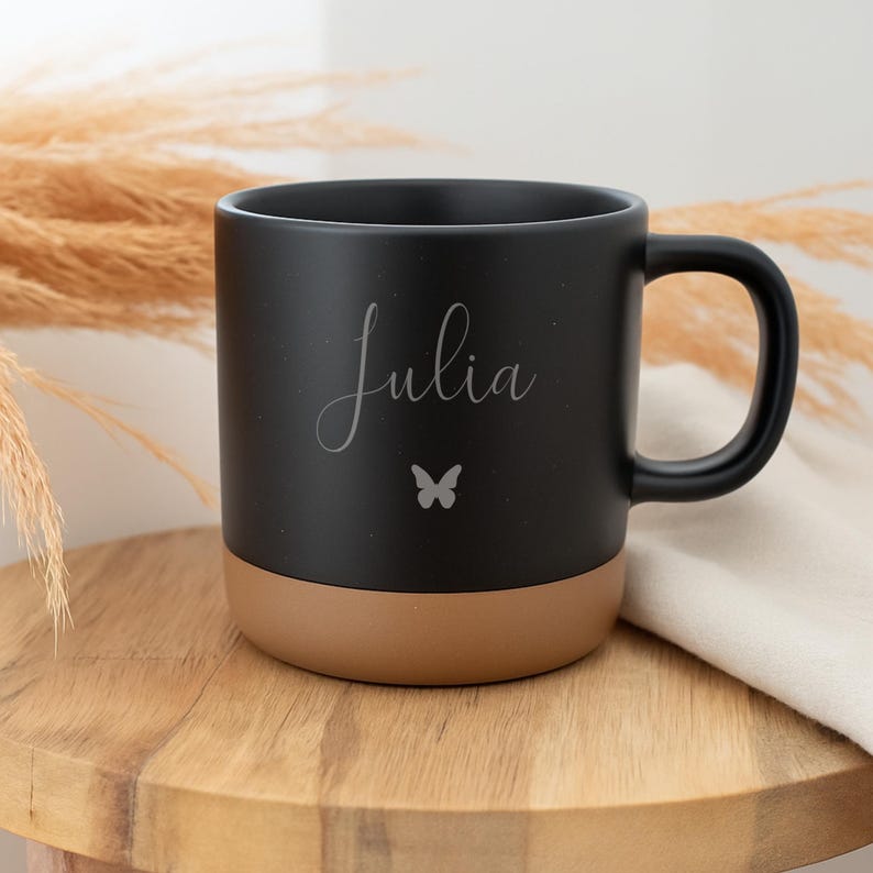 May include: Black ceramic mug with a brown base, featuring the name "Julia" in cursive script and a small butterfly. The mug has a curved handle and is set on a wooden surface.