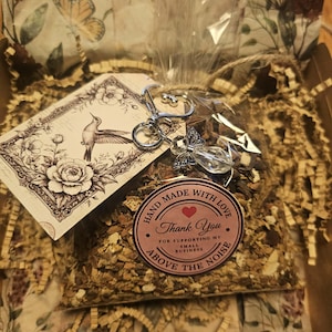 May include: A clear plastic bag filled with dried botanical material, featuring a silver-toned angel charm keychain and a rectangular tag with a hummingbird and floral illustration. A round pink sticker reads: Hand Made With Love Thank You For Supporting My Small Business Above The Noise.
