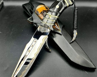 May include: A custom-made knife with a polished steel blade and a black handle with gold accents. The knife is in a black leather sheath with a decorative skull design. A black lanyard is attached to the handle.