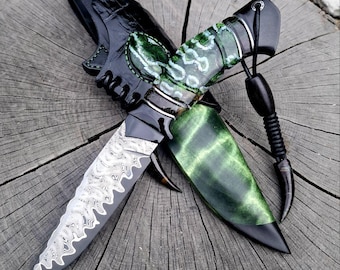 May include: A handcrafted knife with a patterned blade and a green and black handle. The knife is paired with a black leather sheath. The blade has a unique design, and the handle features a decorative pattern. The knife is displayed on a wooden surface.
