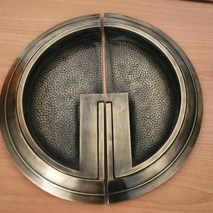 Puede incluir: Un tirador de puerta redondo de latón antiguo con acabado martillado. El tirador está dividido en dos mitades, cada una con un diseño rectangular en relieve.