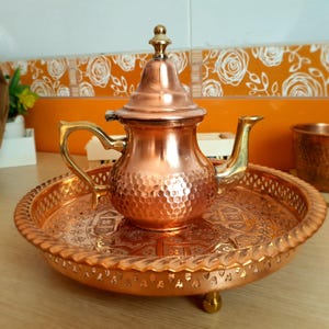 Peut inclure: Une théière en cuivre avec une texture martelée, une poignée et un bec dorés, et un couvercle décoratif. La théière repose sur un plateau en cuivre assorti avec un motif orné et un bord tressé. Le plateau a de petits pieds dorés.