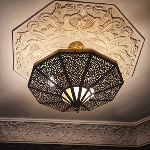 May include: A black metal ceiling light fixture with intricate cutouts and a white frosted glass shade. The light fixture is hanging from a white ceiling with ornate plasterwork.