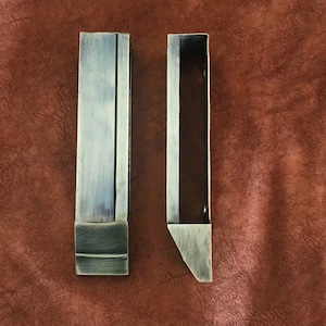 May include: Two rectangular, antique-finished metal drawer pulls. One is closed, the other is open, revealing a hollow interior. The pulls are a muted silver color with darker accents, set against a textured, reddish-brown background.