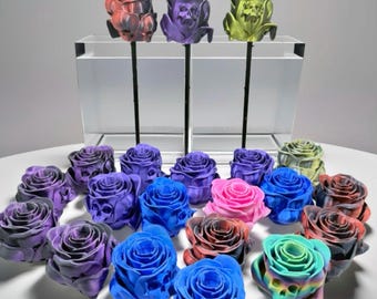 May include: A collection of 15 artificial roses in various colors, including black, purple, pink, blue, green, and red. The roses are arranged in a circle on a white surface. Some of the roses have a skull design.