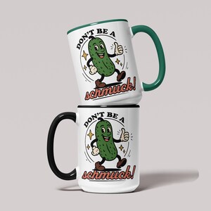 May include: Two white ceramic mugs stacked on top of each other. The top mug has a green handle and rim, and the bottom mug has a black handle and rim. Each mug features a cartoon pickle character with the text "DON'T BE A schmuck!"