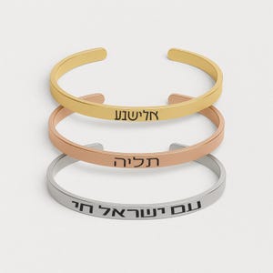 May include: Three open cuff bracelets in gold, rose gold, and silver. Each bracelet has Hebrew text engraved on the front. The gold bracelet reads "אלישבע", the rose gold bracelet reads "תליה", and the silver bracelet reads "עם ישראל חי".