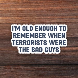 May include: A white sticker with the text "I'M OLD ENOUGH TO REMEMBER WHEN TERRORISTS WERE THE BAD GUYS" in dark blue, set against a dark brown wood grain background. The sticker is rectangular with rounded corners.