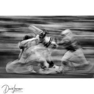 May include: Black and white photograph of a football game in motion. The image captures the blurred action of players in helmets and uniforms, creating a sense of speed and dynamism. The signature "David Lawrence Photographs" is in the bottom left corner.