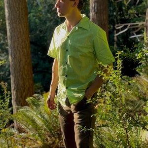 Puede incluir: Un hombre con una camisa de manga corta verde con estampado y cuello abotonado y pantalones marrones. Lleva una gorra de b&eacute;isbol con estampado negro y azul. El hombre est&aacute; de pie en un entorno boscoso.