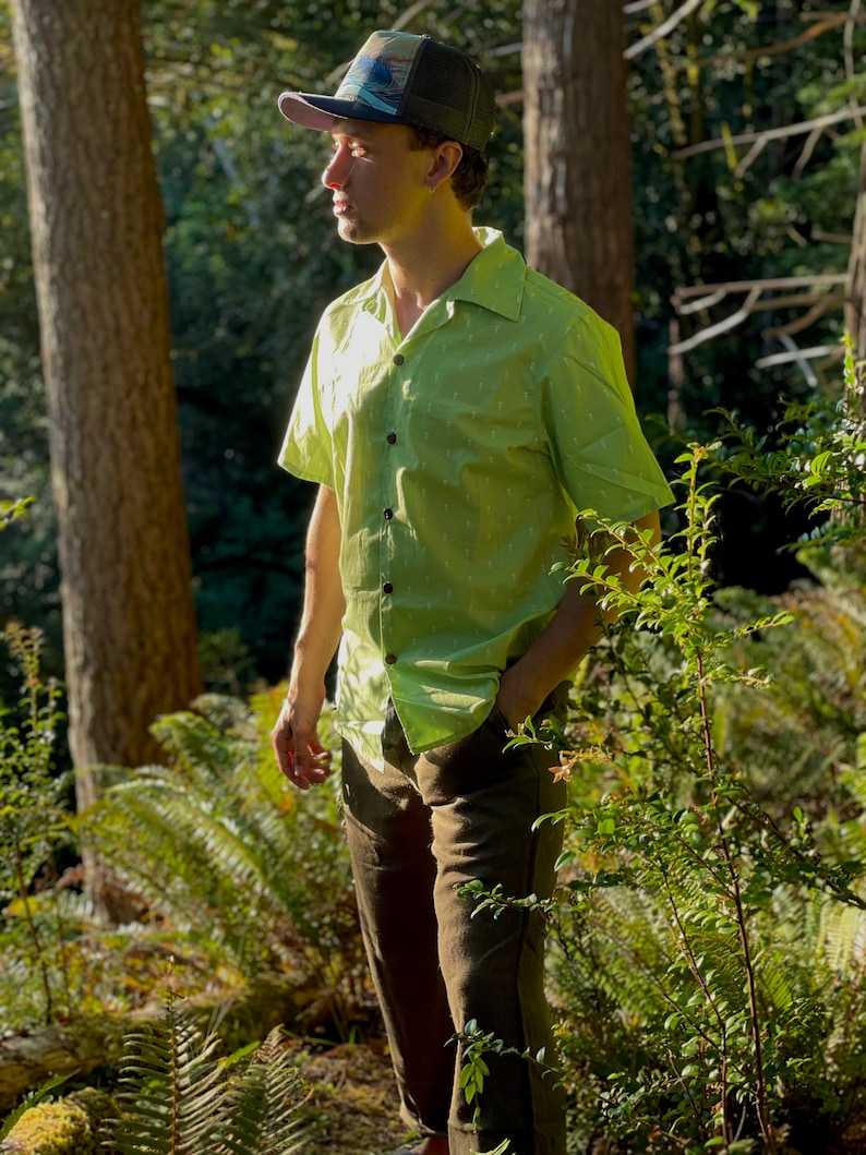 Puede incluir: Un hombre con una camisa de manga corta verde con estampado y cuello abotonado y pantalones marrones. Lleva una gorra de b&eacute;isbol con estampado negro y azul. El hombre est&aacute; de pie en un entorno boscoso.