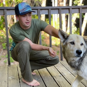 Puede incluir: Un hombre con una camisa verde y una gorra de b&eacute;isbol con estampado azul y blanco est&aacute; en cuclillas en una plataforma de madera. Est&aacute; mirando a un perro que corre hacia &eacute;l. El perro es una mezcla de gris y blanco.