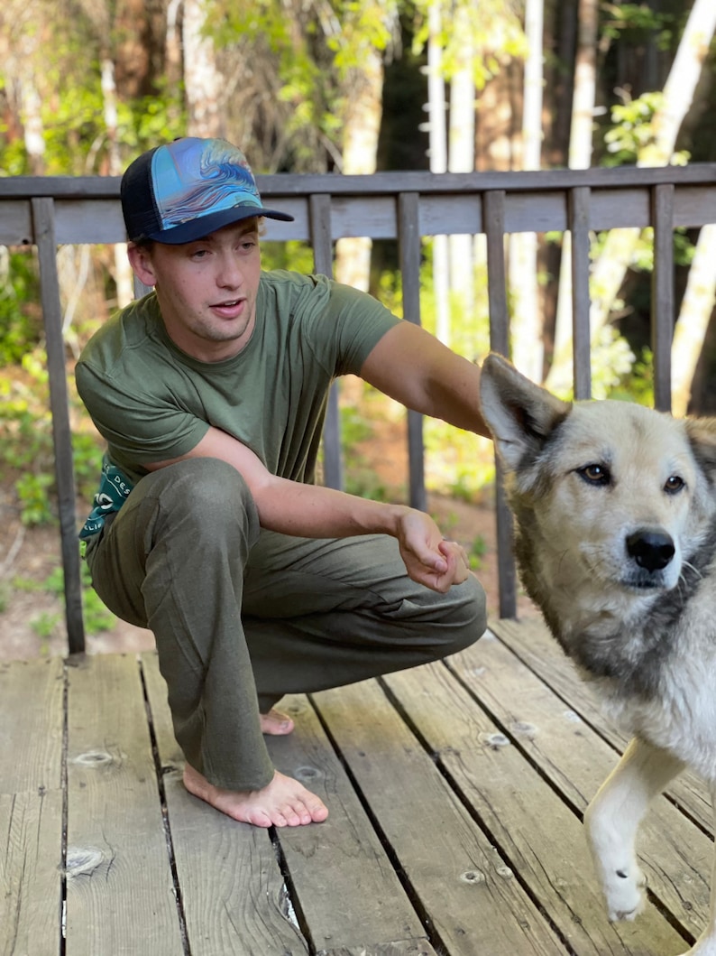 Puede incluir: Un hombre con una camisa verde y una gorra de b&eacute;isbol con estampado azul y blanco est&aacute; en cuclillas en una plataforma de madera. Est&aacute; mirando a un perro que corre hacia &eacute;l. El perro es una mezcla de gris y blanco.