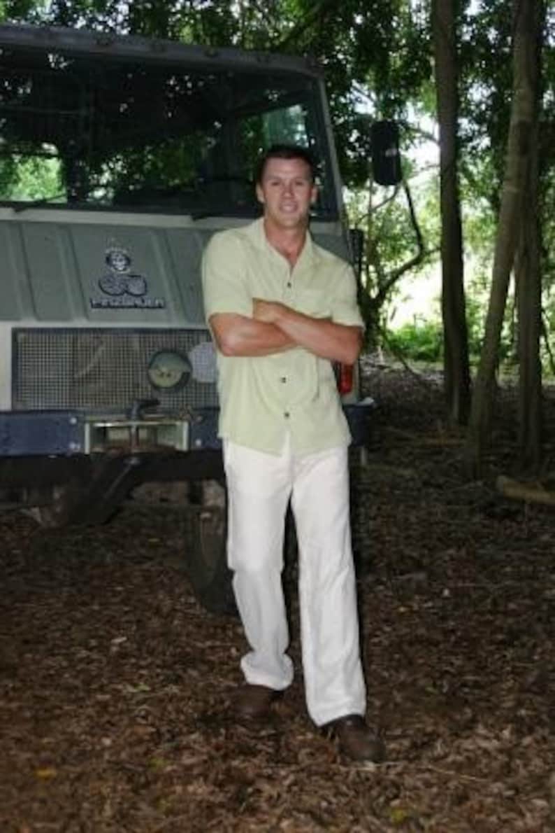 Puede incluir: Un hombre con una camisa de manga corta verde claro y pantalones blancos est&aacute; de pie frente a un cami&oacute;n grande de color gris. El cami&oacute;n tiene una parrilla negra y un parachoques negro. El hombre sonr&iacute;e y mira a la c&aacute;mara.