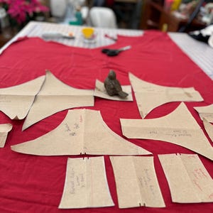 May include: A red fabric surface with multiple brown paper pattern pieces for sewing a garment. The pattern pieces are labeled with handwritten instructions, including "Bust Dart", "Back Neck", "Shoulder Piece", "Bust", "Back", and "Front".